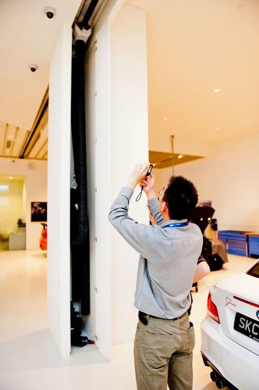 A man taking a photo with a smartphone inside a well-lit indoor space, near a white vehicle with a visible license plate.
