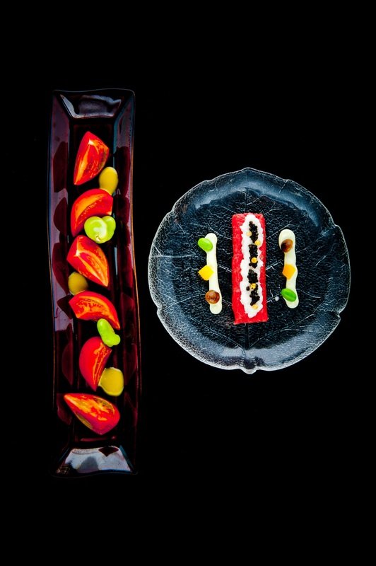 Elegant dessert presentation with a glass rectangular dish containing red and yellow garnishes on the left, and a round black plate with a rectangular red and white layered mousse topped with black and white dots and small green and yellow garnishes,