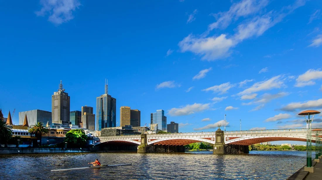Downtown skyscrapers and bridge over river with person kayaking in foreground on a sunny day with blue sky and clouds.