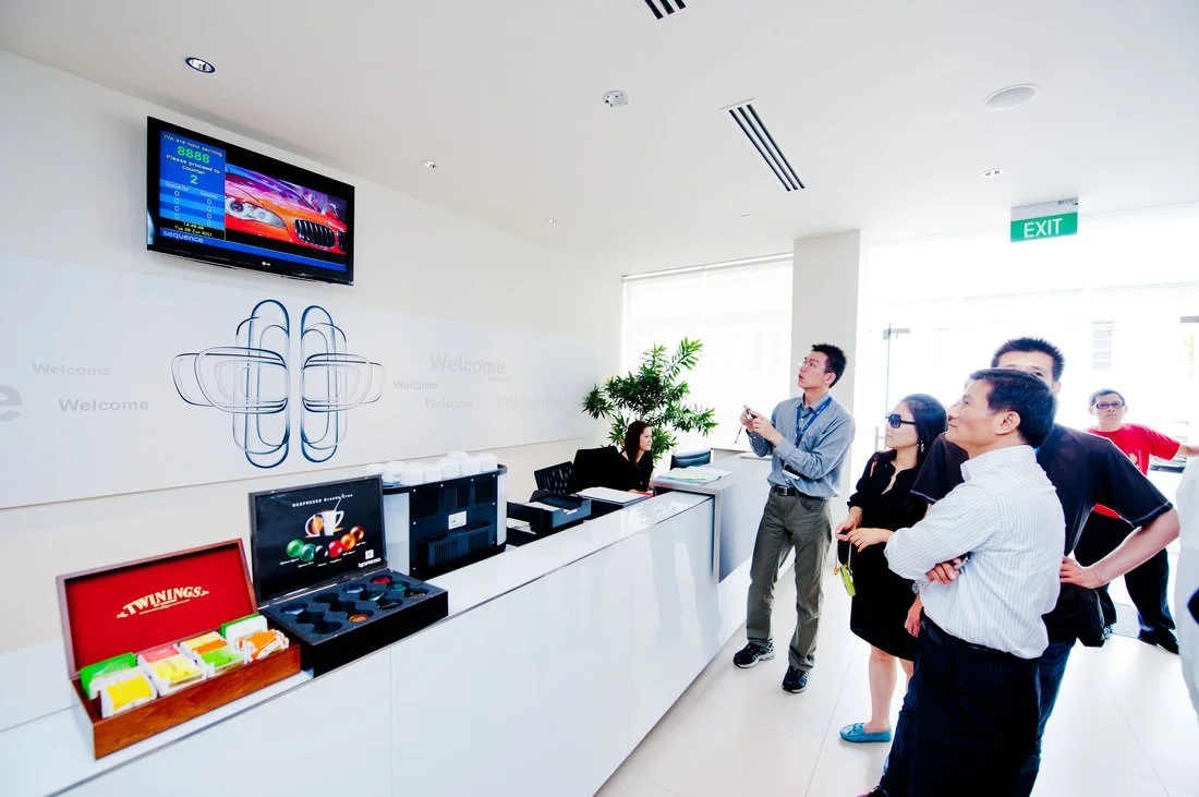Group of people standing near reception desk with snacks and drinks, high ceiling with recessed lighting, TV screen on wall, exit sign, and a large window letting in natural light.