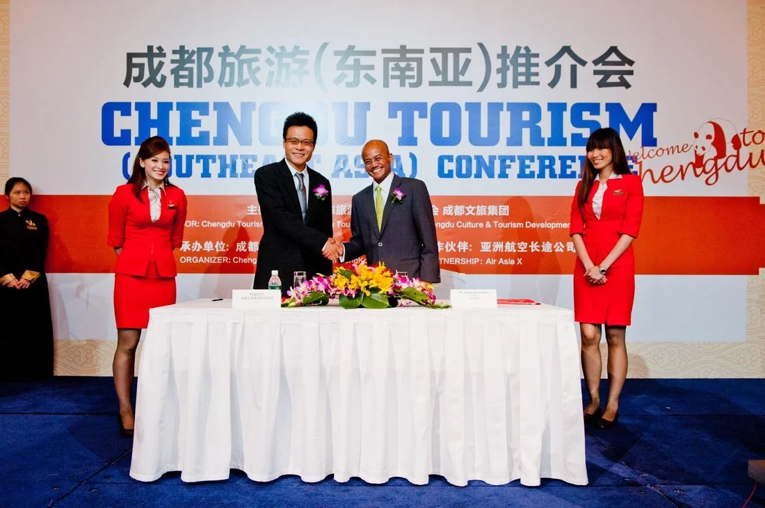 Two men in suits shaking hands in front of a table with flowers at a Chengdu tourism conference in China, with three women in red uniforms standing beside them.