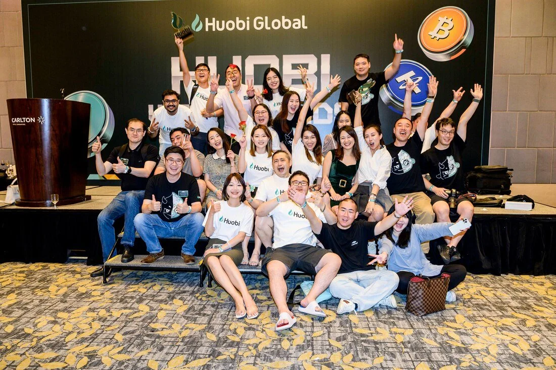 Group of smiling people celebrating at a conference, with a backdrop displaying UBI and cryptocurrency symbols, some wearing Huobi t-shirts.