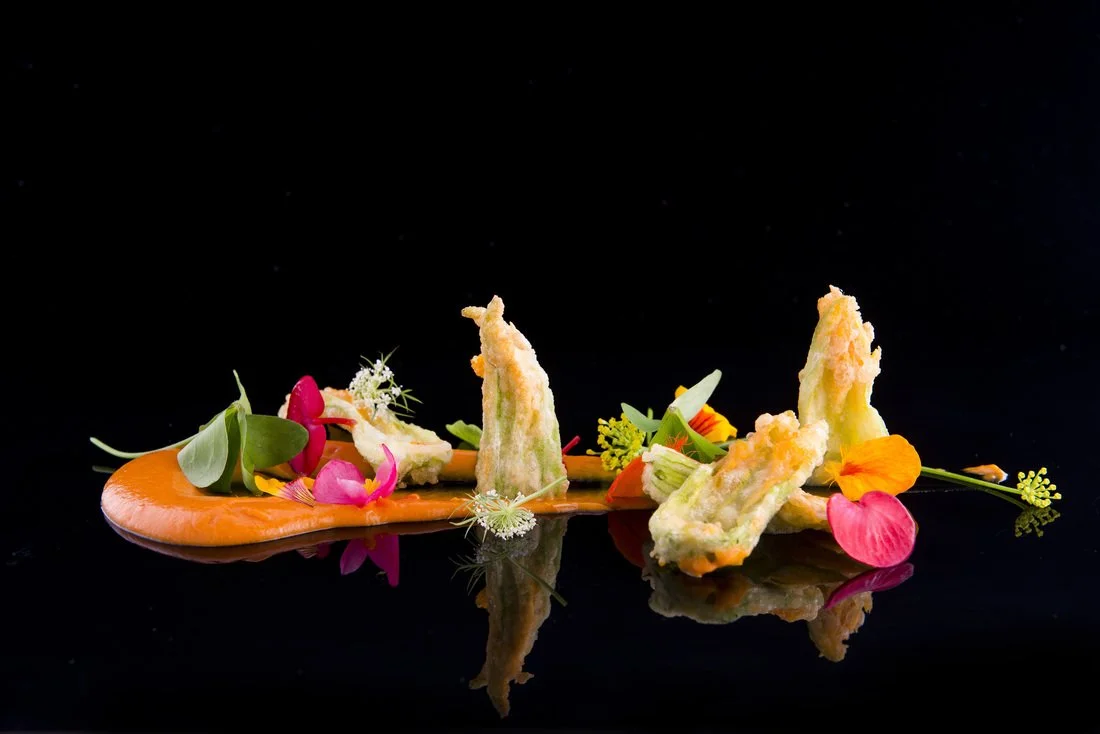 Gourmet dish with tempura vegetables, colorful edible flowers, and a drizzle of sauce on a reflective black surface