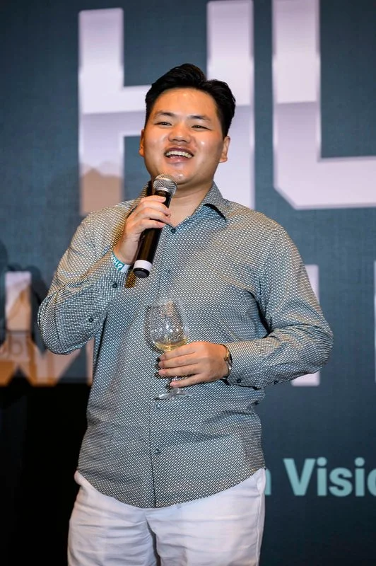 A man smiling and holding a microphone in one hand and a glass of wine in the other, standing in front of a dark background with large white text.