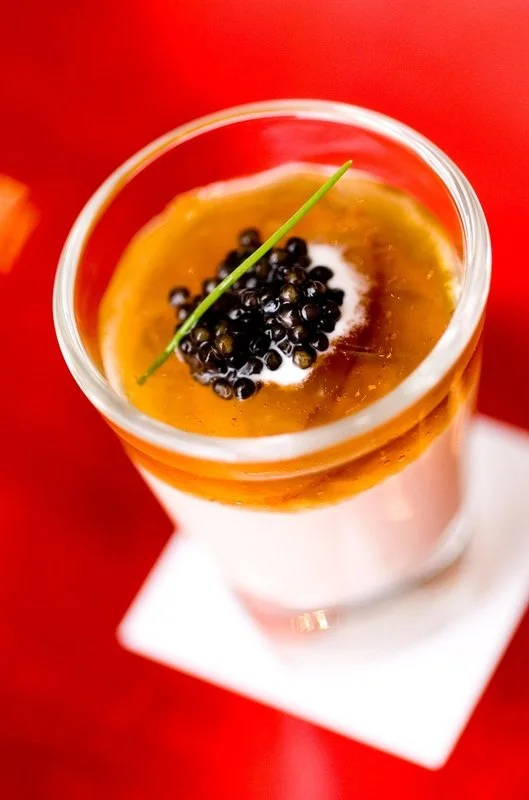 A layered dessert in a clear glass topped with black caviar, a dollop of cream, and a green garnish on a white napkin, on a red surface.