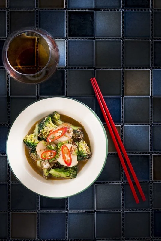 A bowl of stir-fried broccoli with red chili peppers on a black tiled table, accompanied by a pair of red chopsticks and a glass of tea.