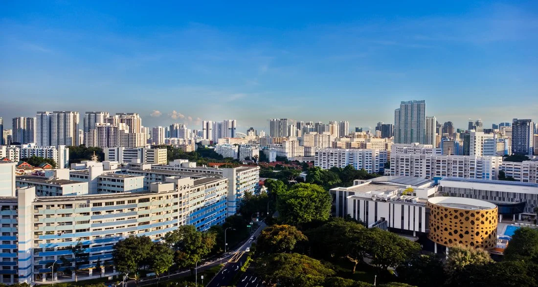 A cityscape with numerous high-rise buildings, some with white and blue exteriors, surrounded by lush green trees, under a blue sky with a few clouds.