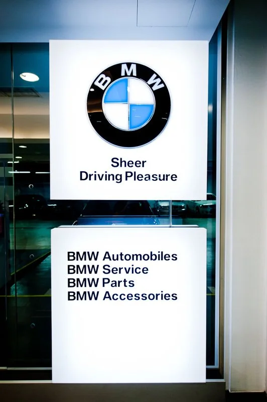 BMW dealership sign with logo and text reading "Sheer Driving Pleasure" and listing services: BMW Automobiles, BMW Service, BMW Parts, BMW Accessories.