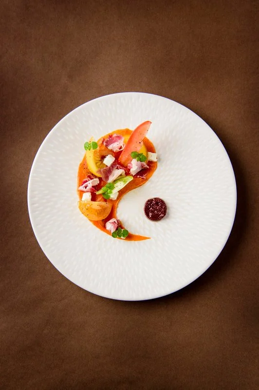Elegant plated dish featuring a colorful array of vegetables and edible flowers arranged artistically on a white textured plate with a small dollop of sauce or puree.