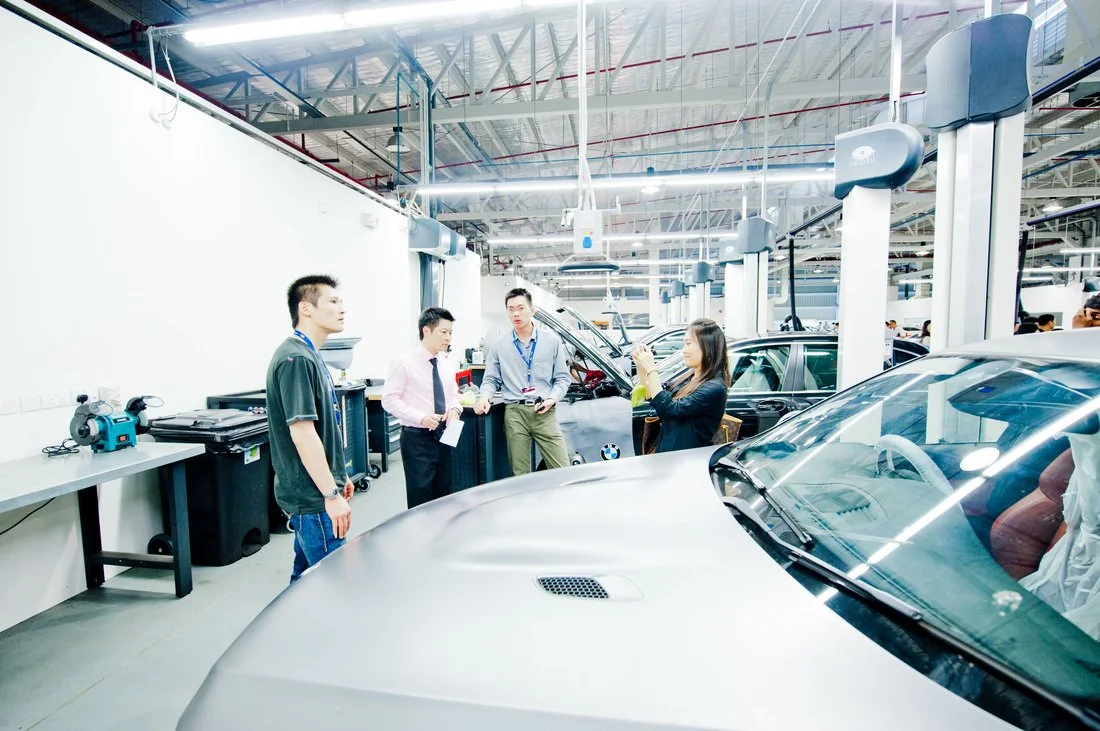 Group of people at an automotive manufacturing or repair plant with cars and equipment in the background.