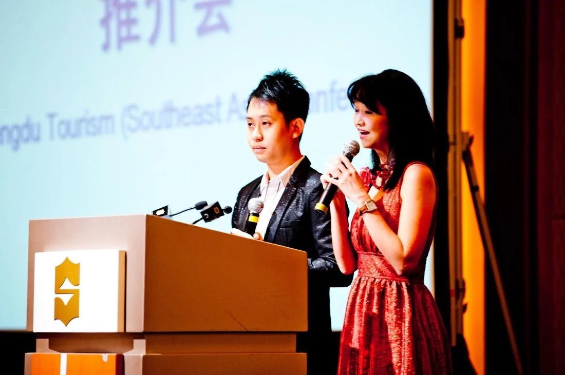 Two young women speaking at a podium with microphones during an event, with a large screen in the background displaying text about Southeast Asia tourism.