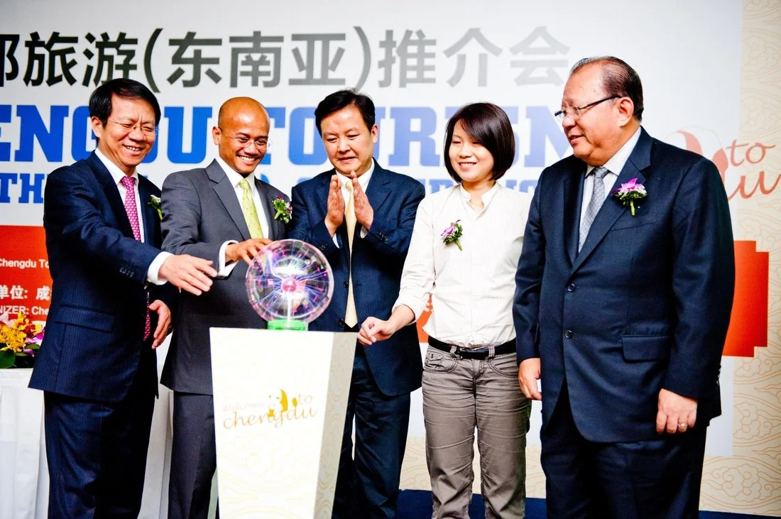 Five businesspeople, dressed in formal suits and wearing flower lapel pins, participate in a ceremonial event with a glowing orb on a pedestal. They are smiling and engaging with the orb in front of a backdrop with Chinese and English text, suggestin