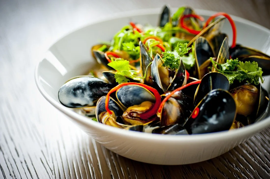 Bowl of steamed mussels garnished with red chili peppers and fresh cilantro on a wooden surface.