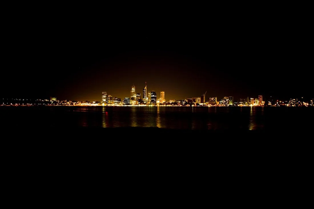 Nighttime city skyline reflecting on water with illuminated skyscrapers and dark sky.
