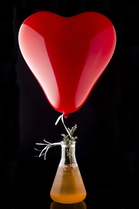 A red heart-shaped balloon tied to a glass container with a yellowish liquid, decorated with small white flowers and white ribbons.