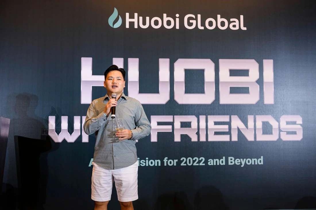 A man speaking into a microphone at a Huobi Global event, standing in front of a backdrop that reads 'Huobi Global' and 'Huobi World Friends' with additional text about a vision for 2022 and beyond.