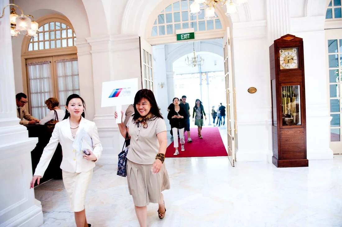 People walking through a grand white lobby with tall windows and a red carpet, including two women smiling and holding up signs, and a clock on a wooden stand.