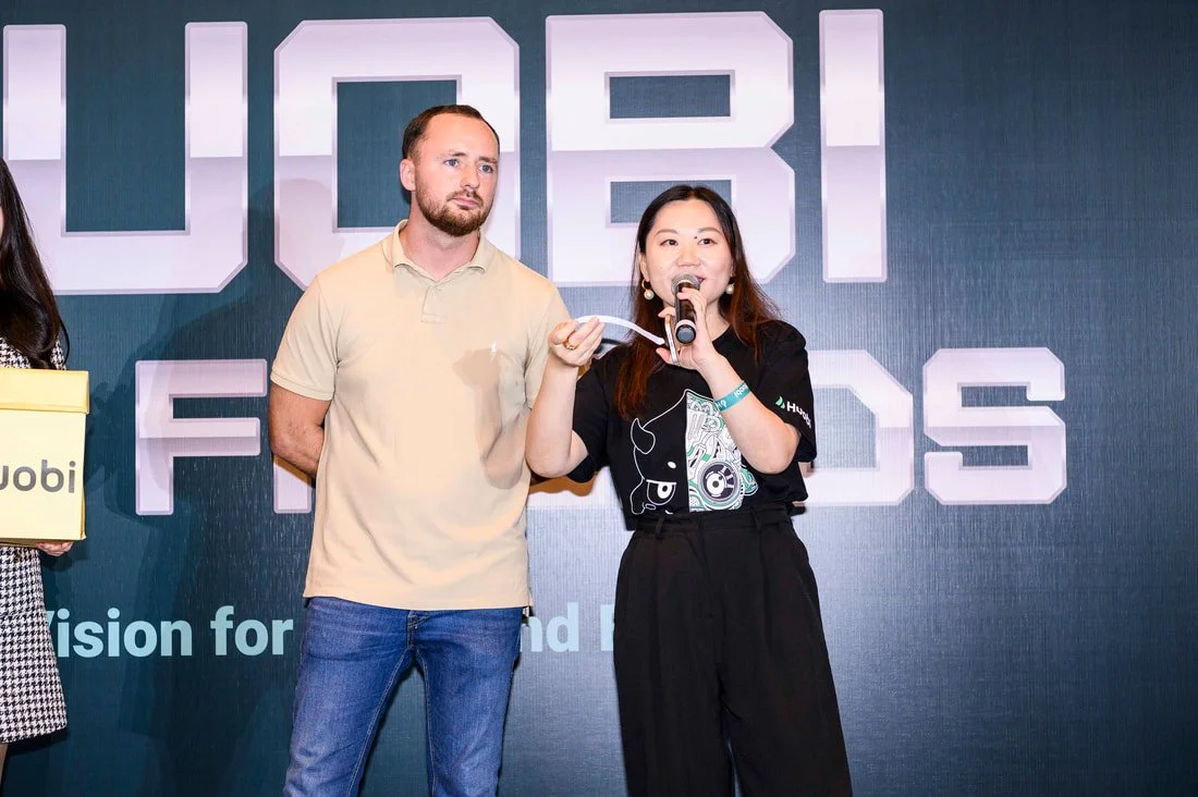 A woman speaking into a microphone on stage, with a man standing beside her, at an event with a large screen behind them displaying the words "L5BI F" and some other indistinct text.