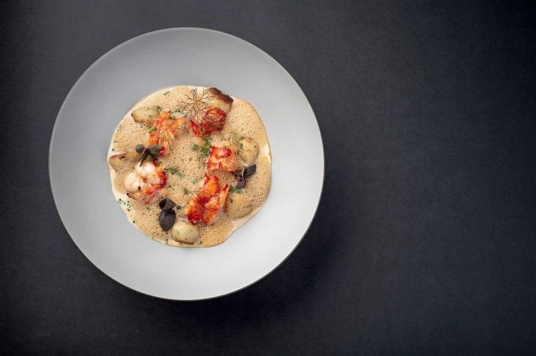 A plate of gourmet lobster and shellfish in a foam sauce, garnished with herbs and microgreens, on a dark surface.