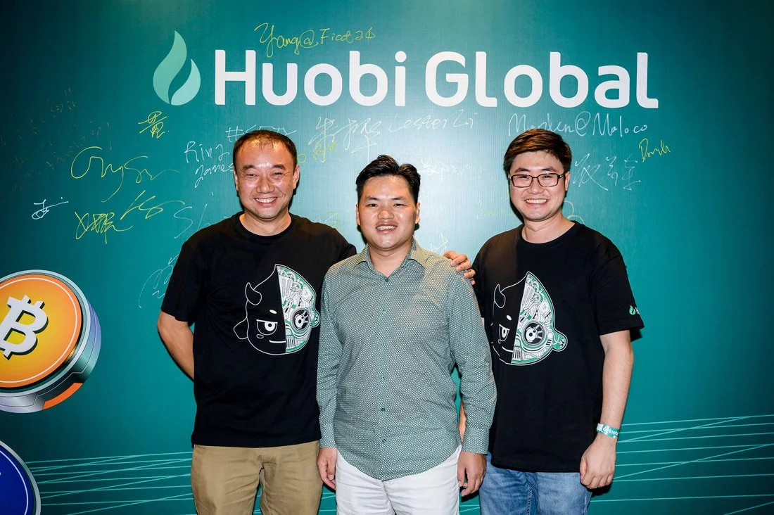 Three men standing together in front of a teal backdrop with the Huobi Global logo. The man in the middle is wearing a green dotted shirt and smiling, with the two men on either side wearing black shirts with a cartoon face design. The backdrop has s