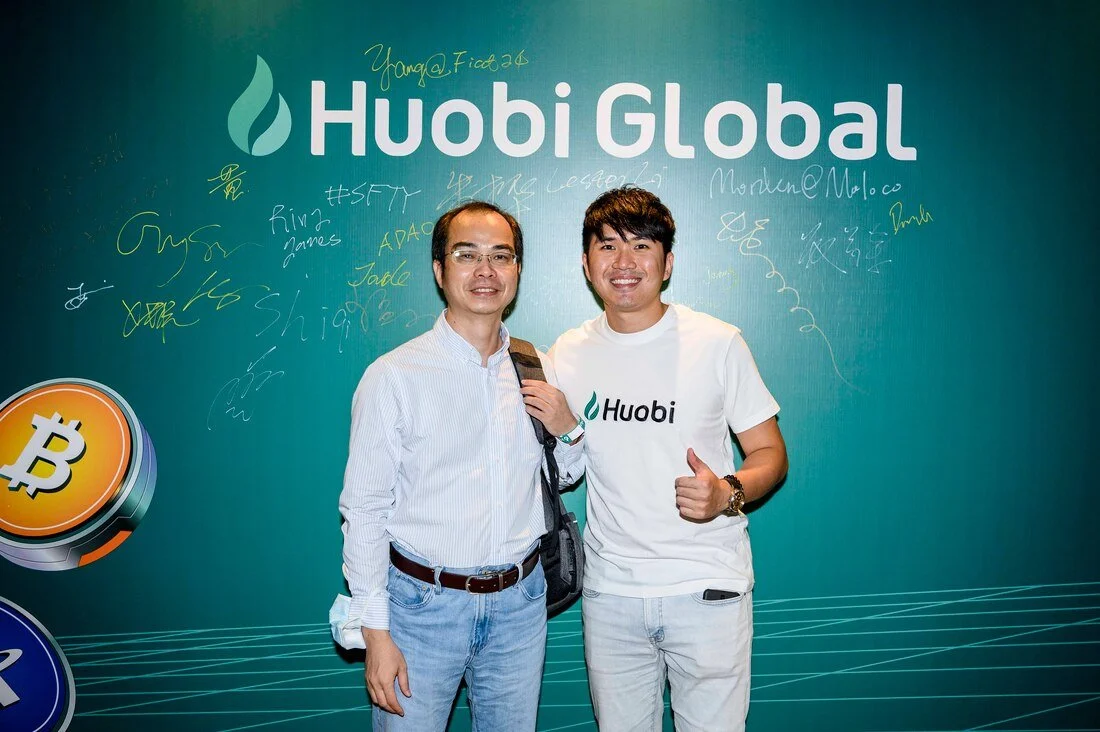 Two men standing in front of a Huobi Global backdrop with handwritten signatures and a Bitcoin graphic.