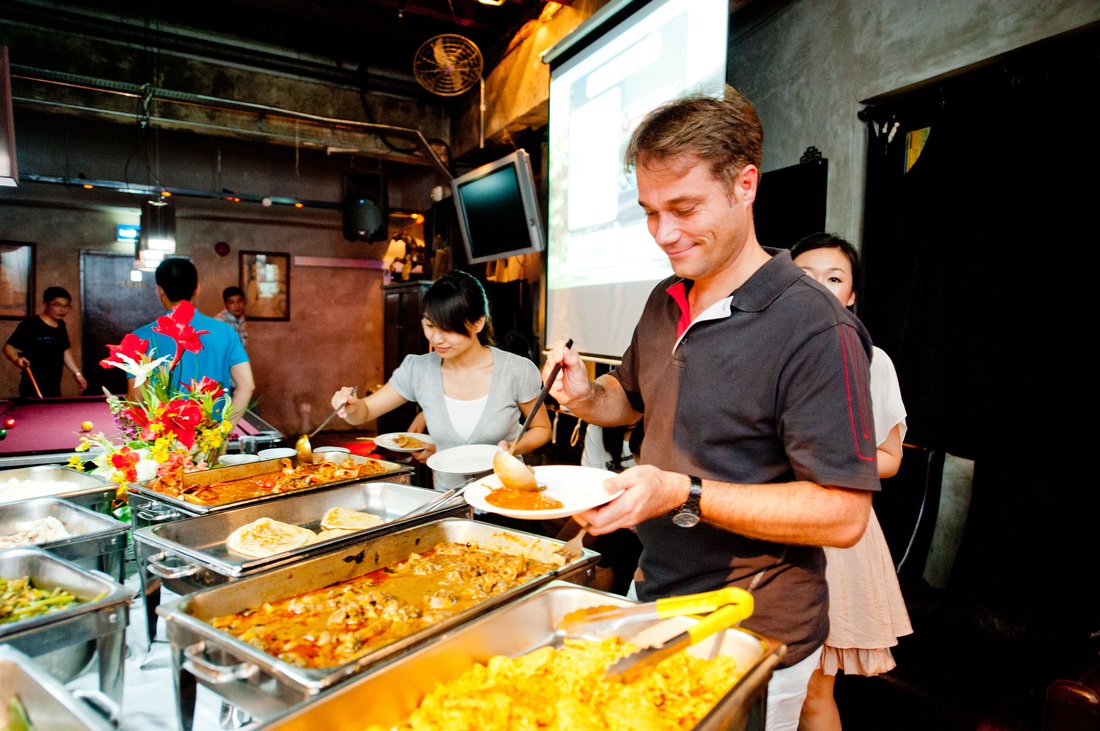 People serving themselves food at a buffet table with various dishes including pizza, curry, and rice in a restaurant or event space.