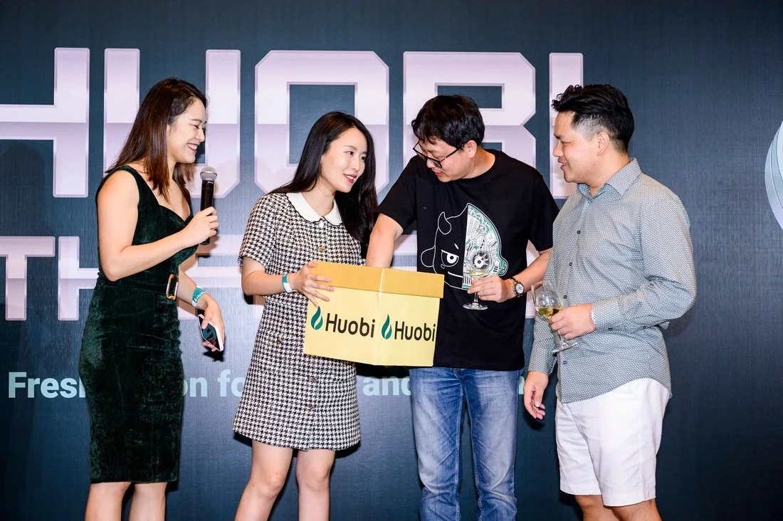 Five people at an event, with one woman holding a microphone, presenting a box labeled 'Huobi' to other three. The group appears to be participating in a presentation or award activity, standing in front of a dark backdrop with partial text.