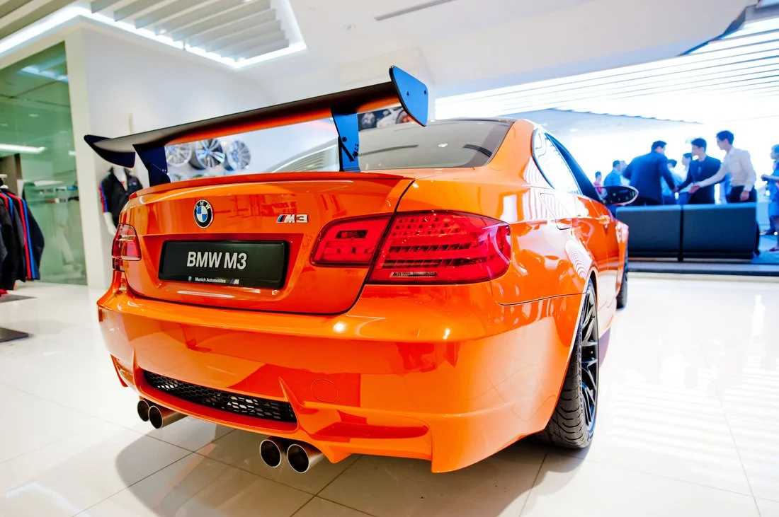 Orange BMW M3 sports car featured in a showroom with a large rear wing spoiler, quad exhaust pipes, and people in the background.