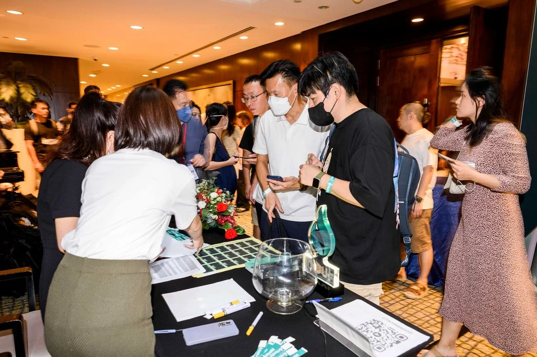 A group of people wearing face masks gather around a table with a raffle drum and papers at an indoor event.
