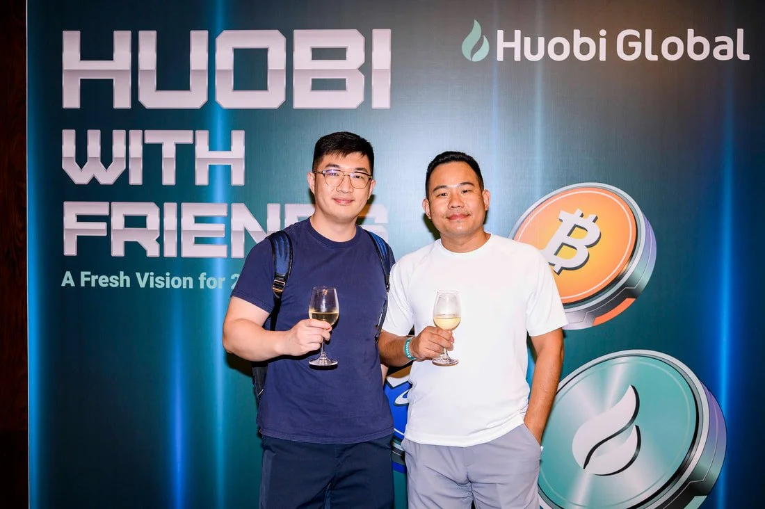 Two men holding glasses of white wine at a Huobi Global event with a blue background displaying the text 'HUOBI WITH FRIENDS'.