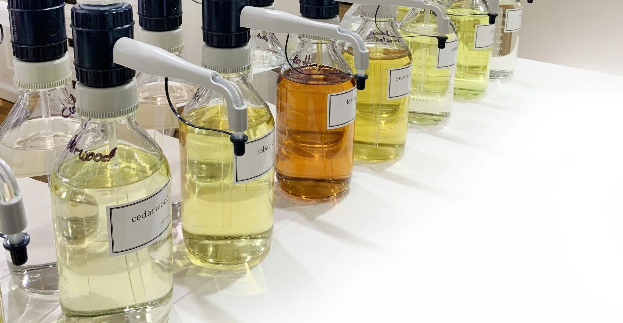 Collection of glass bottles with herbal extracts labeled cedarwood, tobacco, sweet, and others, with spray adapters, on a white table.