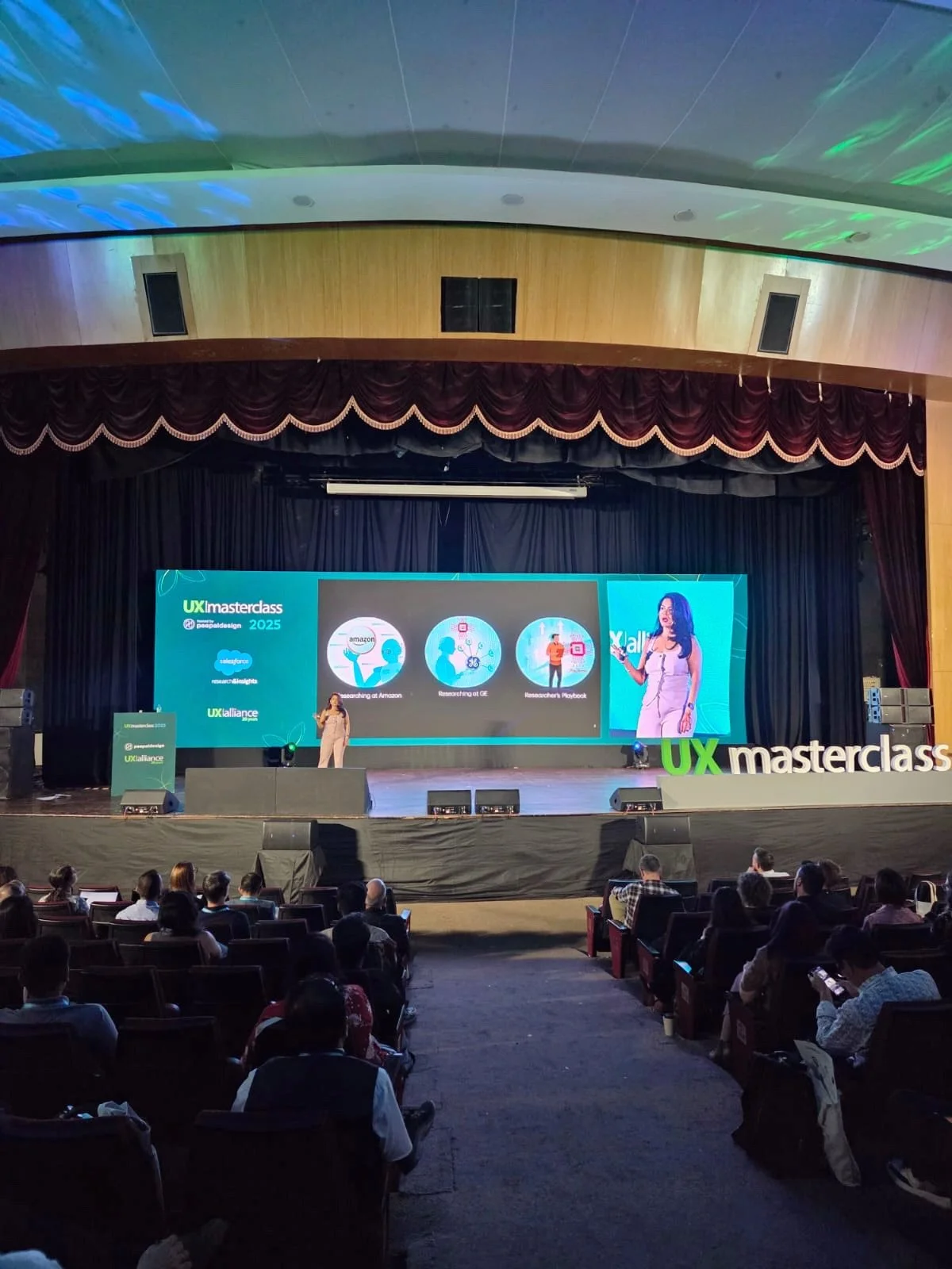 A stage with a large screen displaying the event name 'UX Masterclass 2025' and logos of sponsors, with a speaker presenting to an audience in a theater with red curtains and dark wood accents.