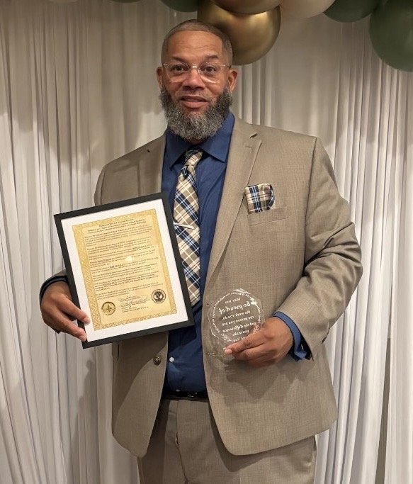 A man in a beige suit, blue shirt, and plaid tie holding a framed certificate in one hand and a clear glass award in the other, standing in front of a white curtain with metallic balloons overhead.