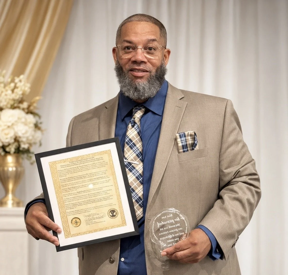 A man with glasses and a gray beard in a beige suit holding a framed certificate and a clear glass award, standing in front of a white curtain with flowers on the side.