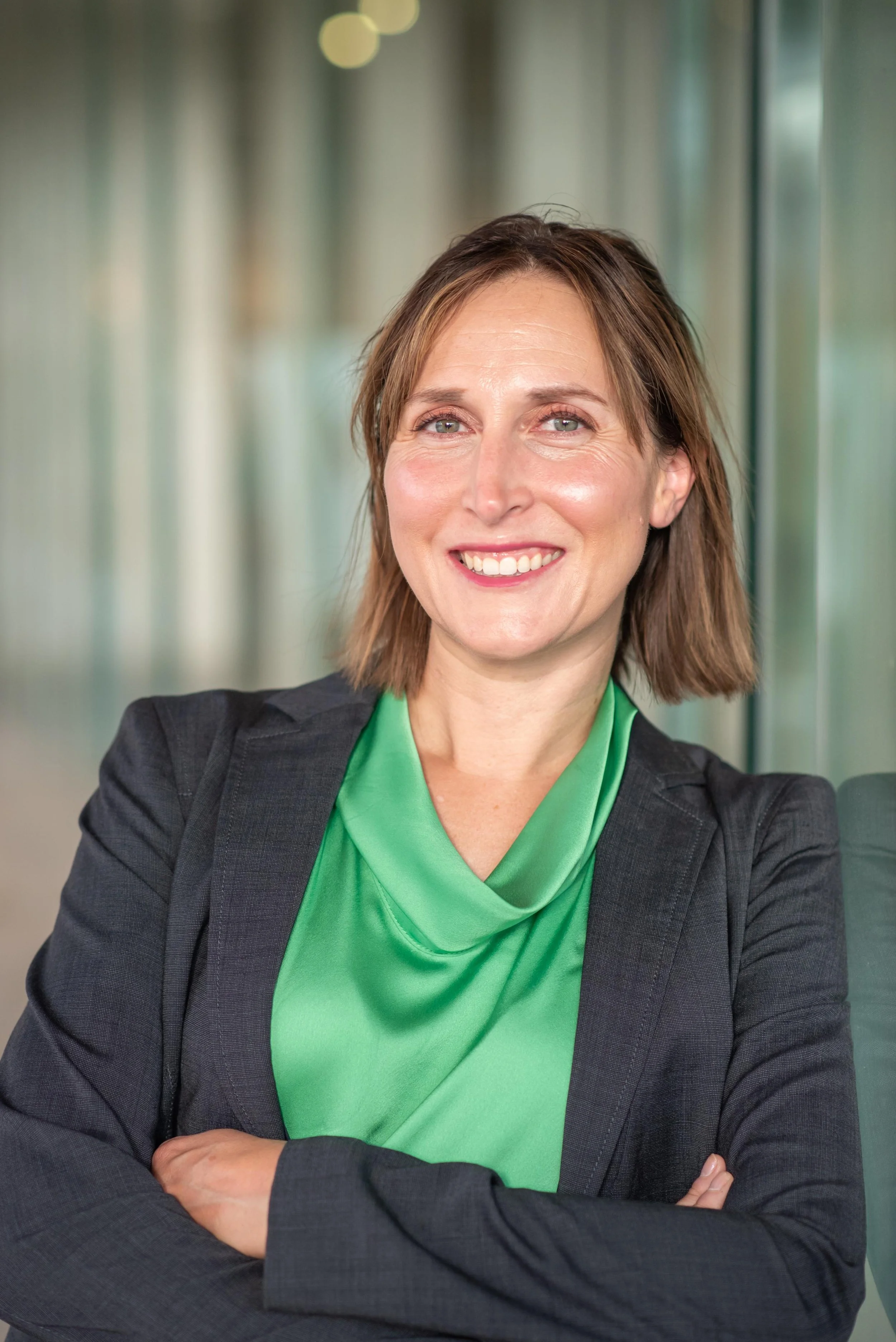 A woman with short brown hair, wearing a black blazer and bright green blouse, arms crossed and smiling in an indoor setting with blurred background.