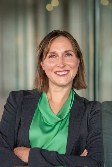 A woman with shoulder-length brown hair, smiling, wearing a black blazer and a green top, standing in front of glass windows with blurred background lights.