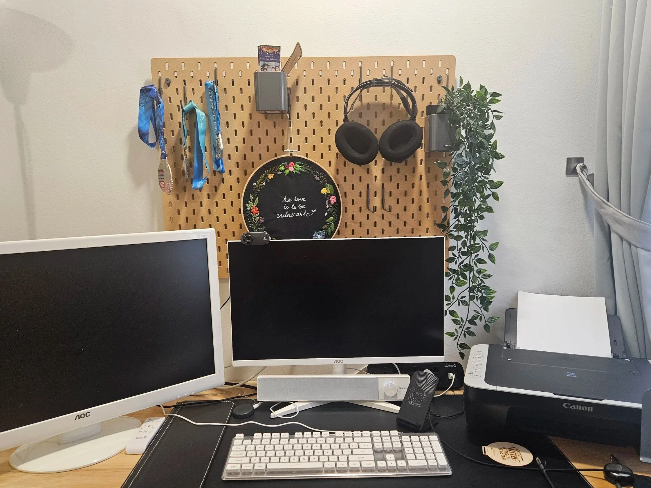 Office desk setup with multiple monitors, a keyboard, a mouse, headphones, a printer, and a folder. A pegboard on the wall behind holds lanyards, a card, and a decorative embroidery hoop with the quote 't to love is to be vulnerable.' There is a potted plant on the right side.