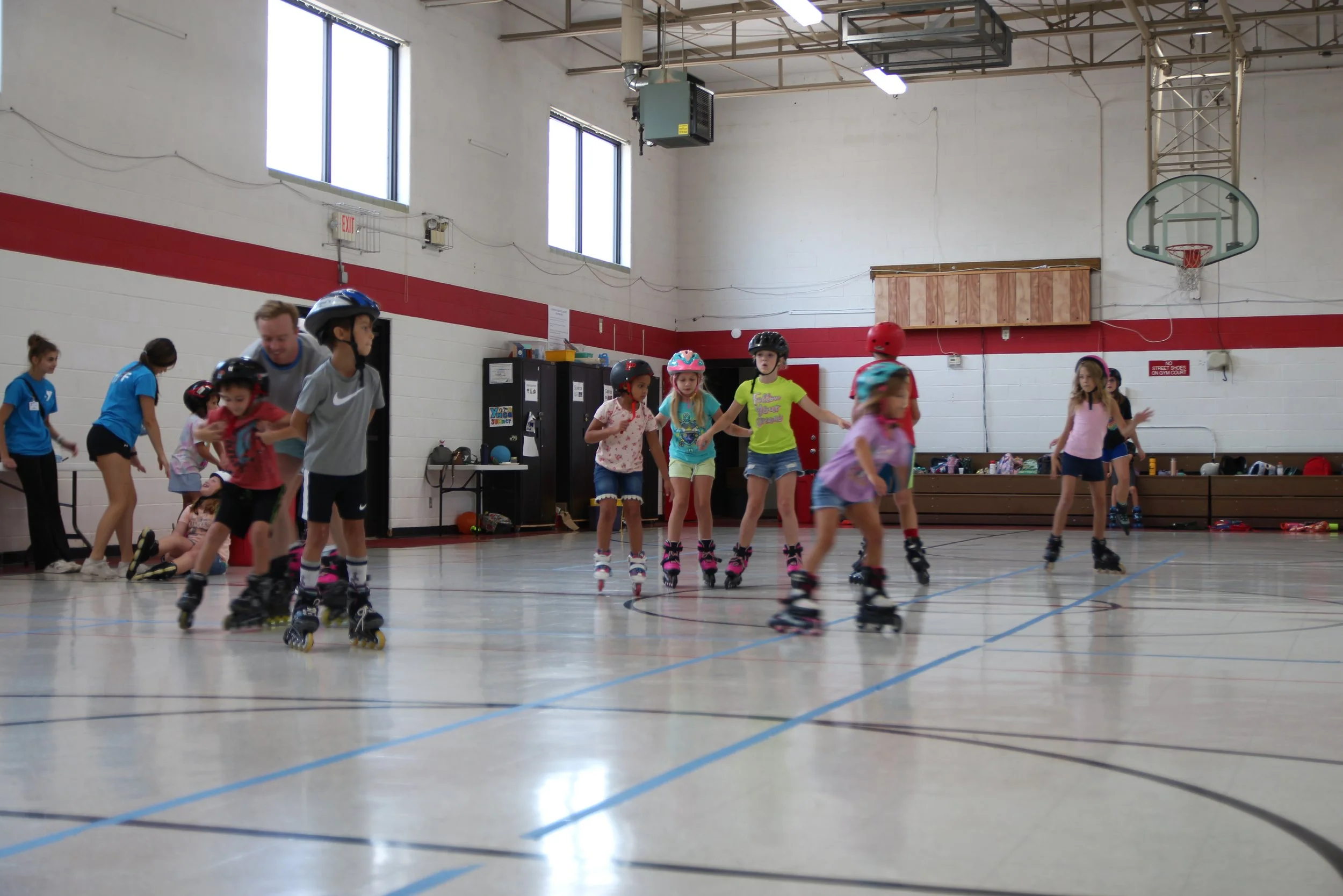 Children wearing helmets roller skating in an indoor gymnasium.