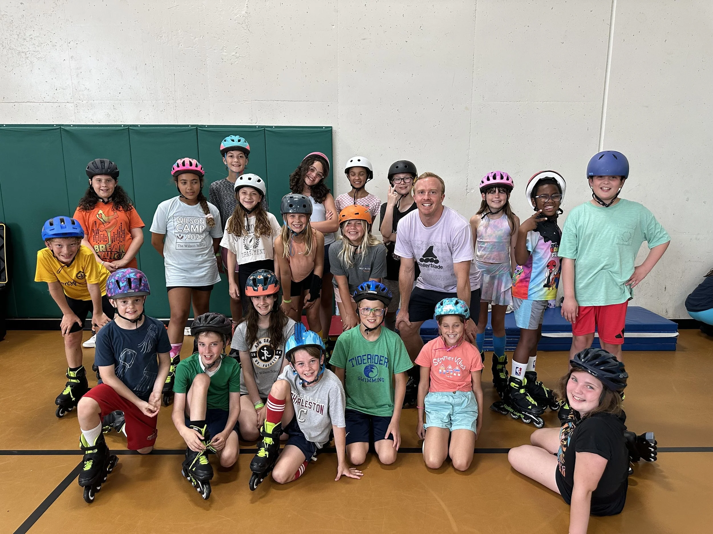 Group of children wearing helmets, some on roller skates, posing indoors on a gym floor with a green padded wall behind them, accompanied by an adult male.