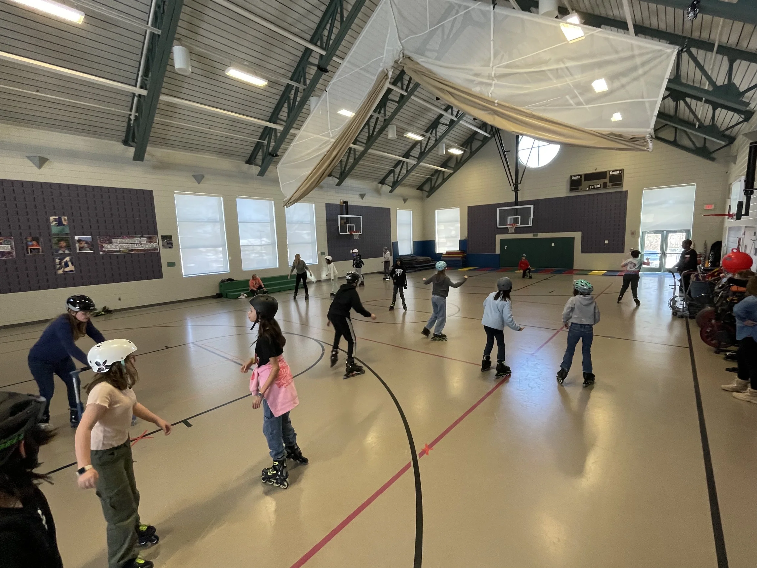 Children and adults roller skating inside a gymnasium with basketball hoops, large windows, and a scoreboard.