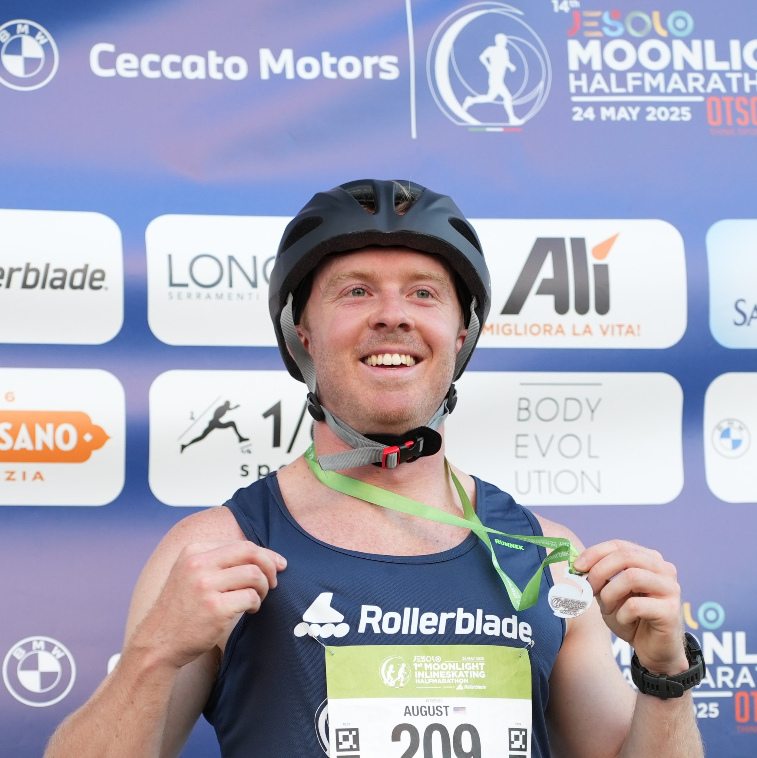 A man wearing a helmet and running gear, smiling, holding and displaying his race medal after participating in the Jesolo Moonlight Half Marathon event.