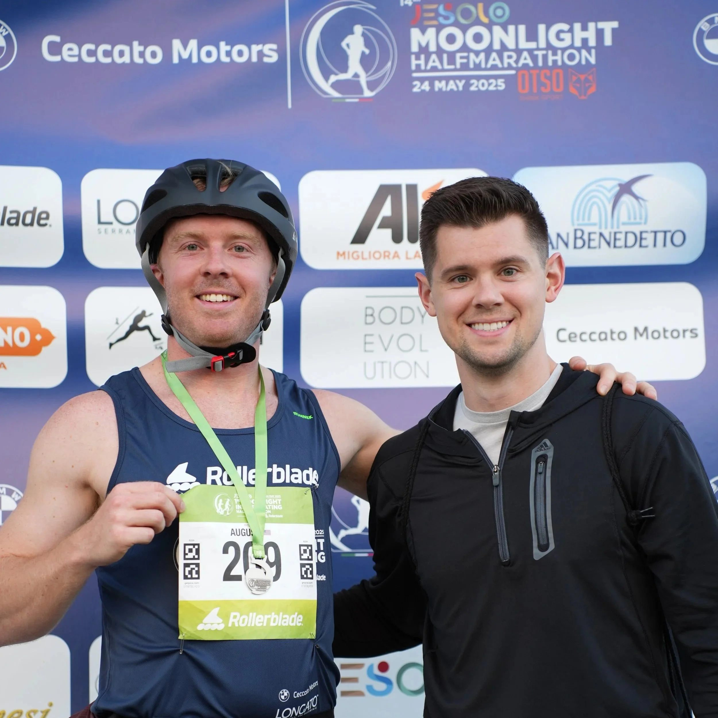 Two men smiling with medals around their necks, standing in front of a blue background with race event logos and text. The man on the left is wearing a helmet and a blue sleeveless shirt, holding a race bib.