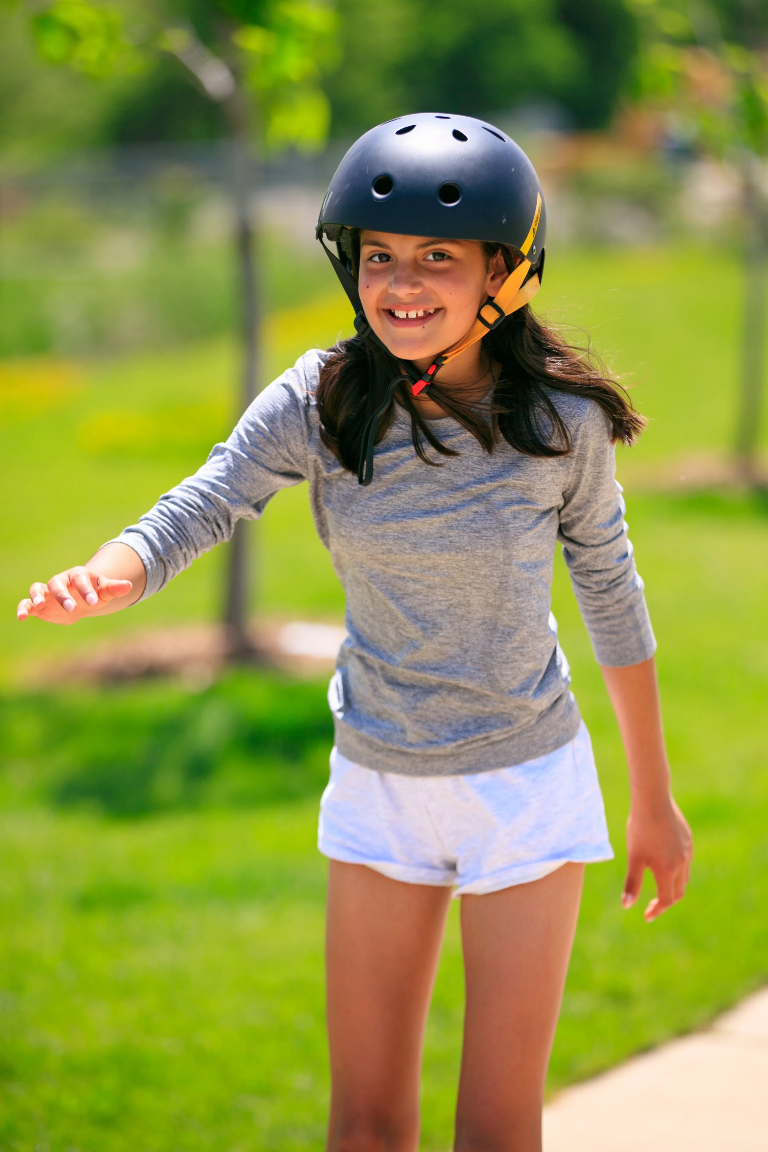 A young girl wearing a black helmet and a gray long-sleeve shirt smiles while outdoors on a sunny day with greenery in the background.