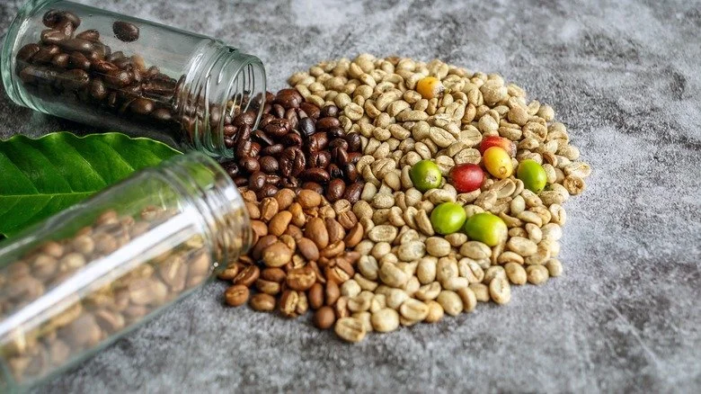 Variety of coffee beans spilling out of glass jars onto a concrete surface, with a green leaf nearby.