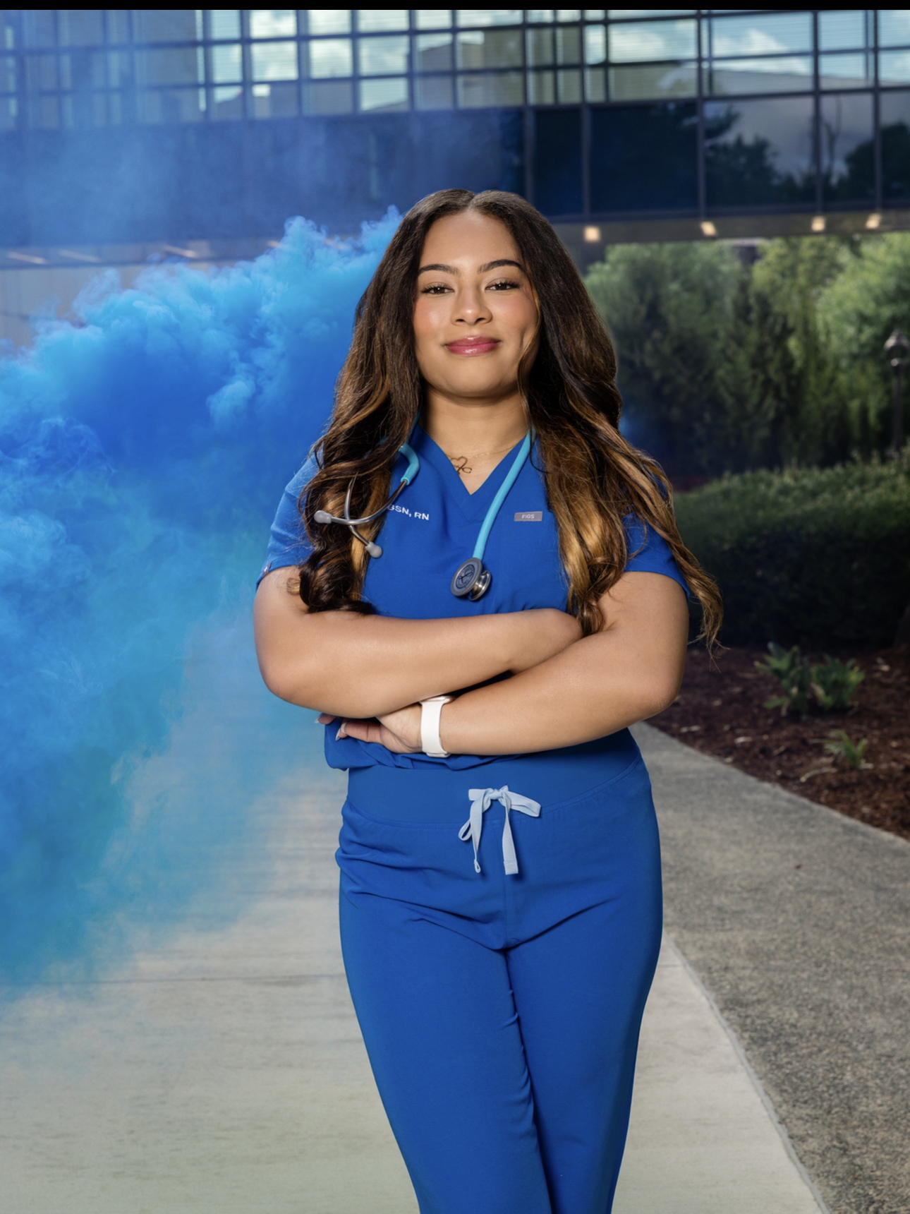 A confident female healthcare professional in blue scrubs and a stethoscope standing outdoors with arms crossed, background with blue smoke, modern building, and greenery.