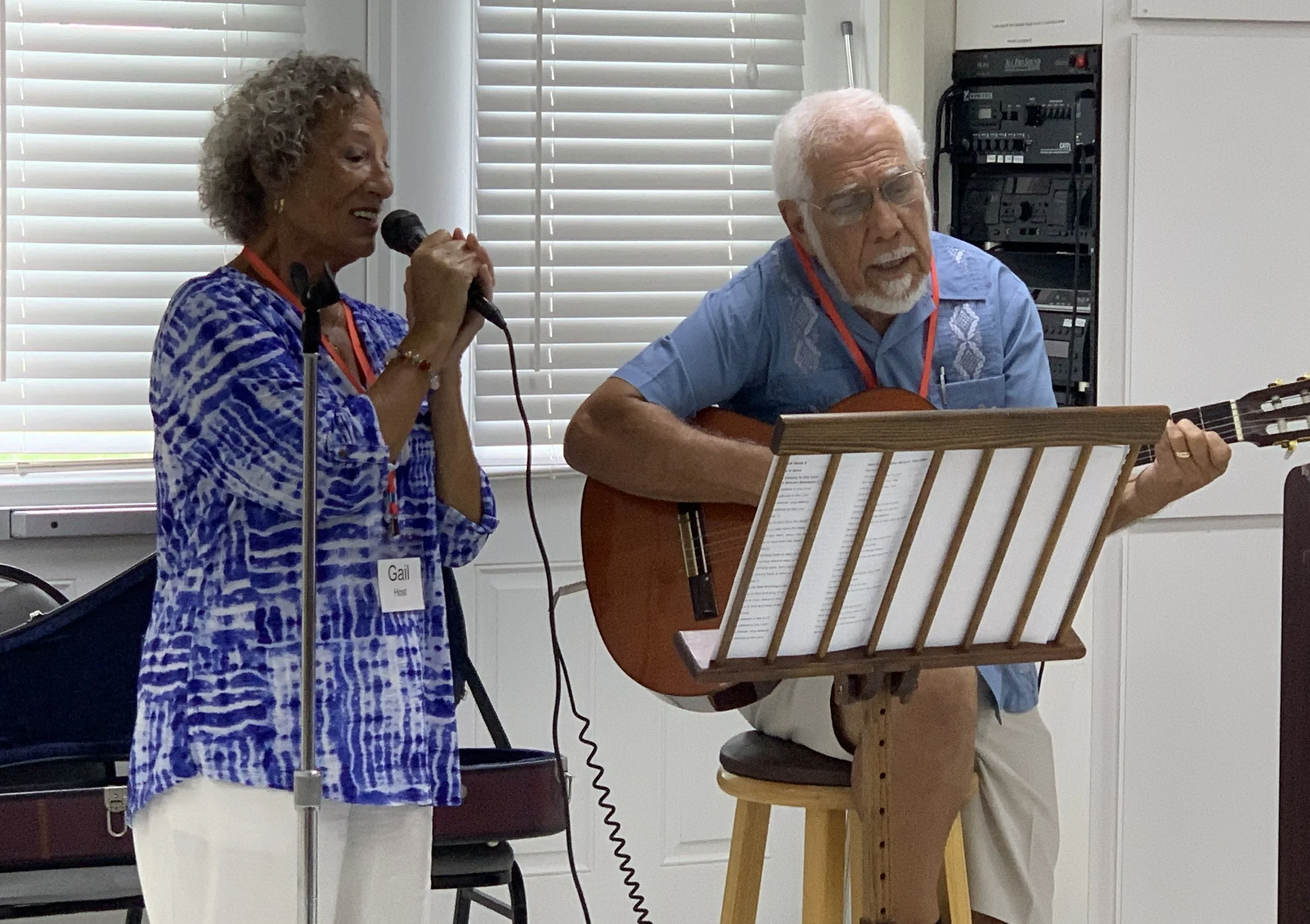 An elderly man playing an acoustic guitar and singing with a woman holding a microphone, during a performance or gathering in a room with white blinds and audio equipment in the background.