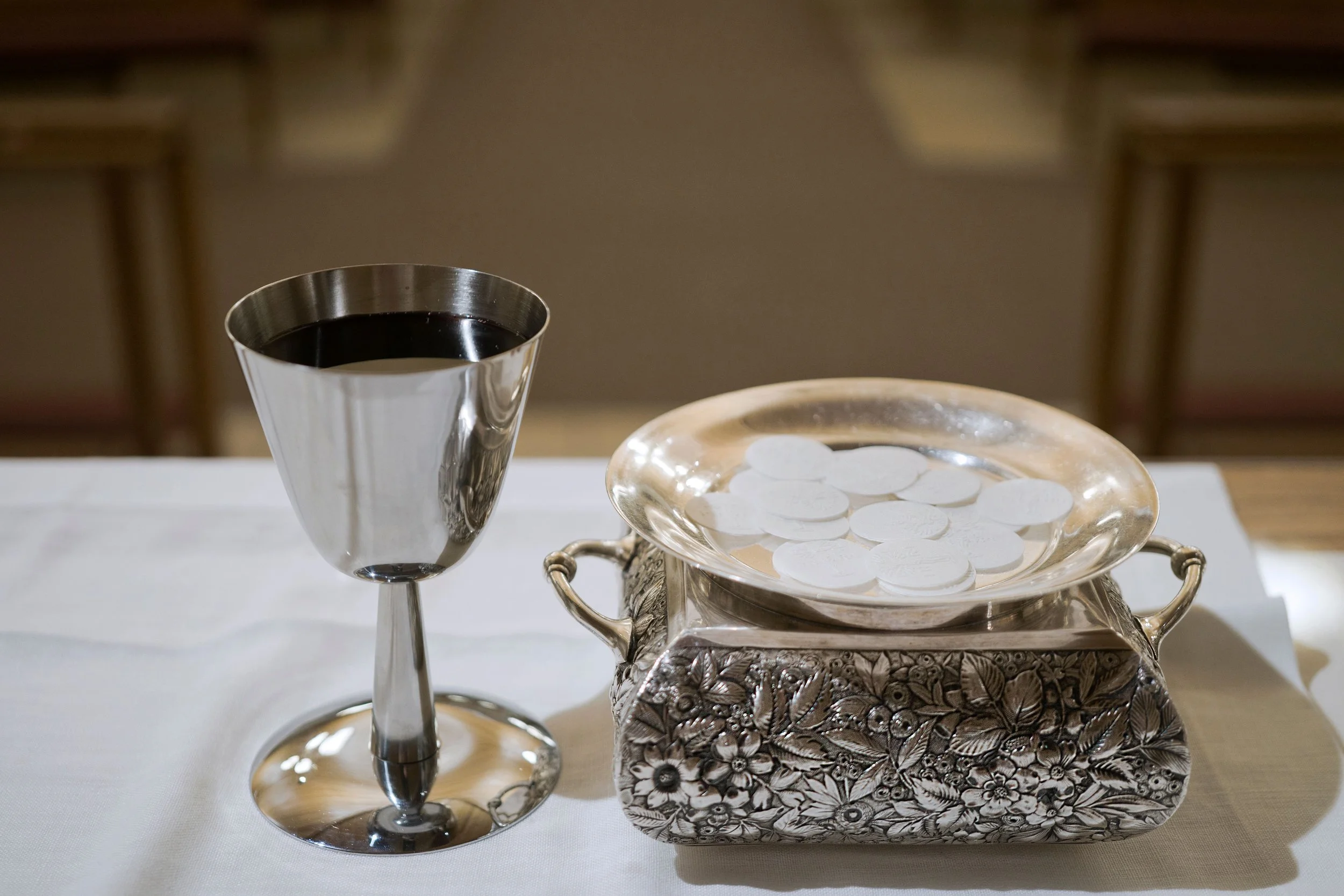 A silver wine glass filled with red wine, a decorative silver tray with white circular tokens, and a ornate silver container with floral patterns on a white tablecloth.