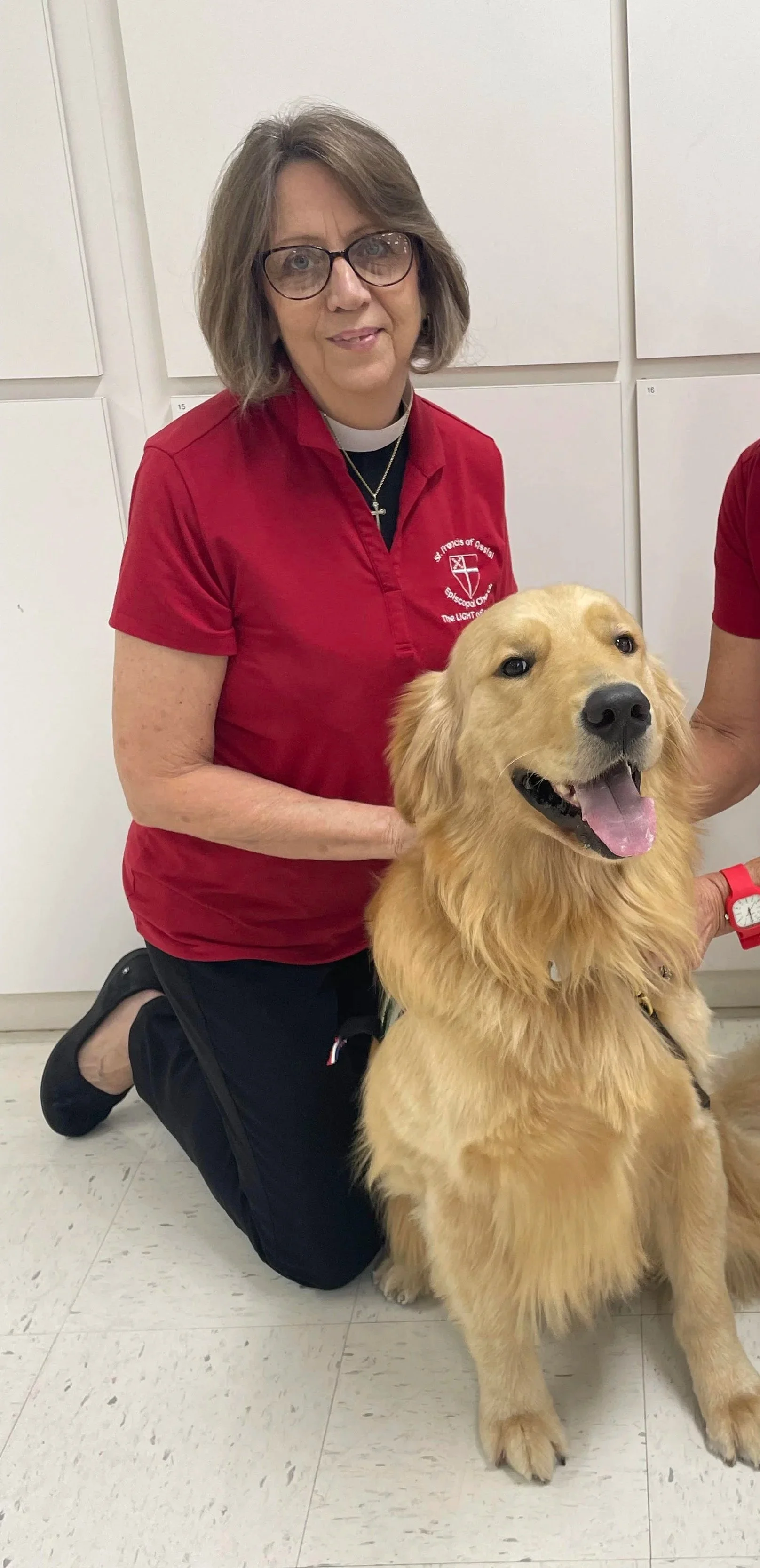 A woman in glasses and a red shirt kneeling next to a golden retriever dog.