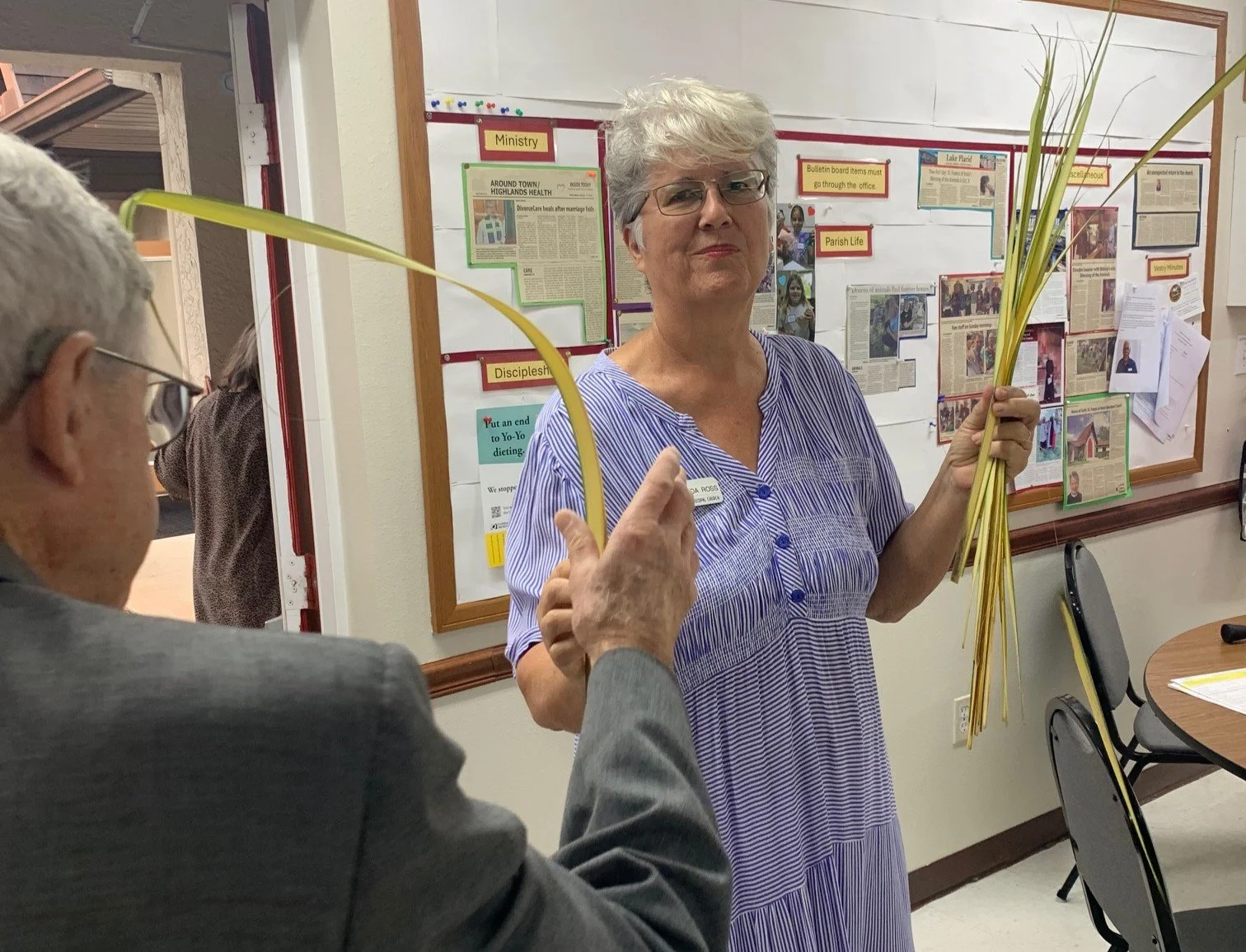 A woman holding a bunch of long yellow palm leaves, standing in front of a bulletin board with articles and signs, while an elderly man with glasses and gray hair gestures towards her.