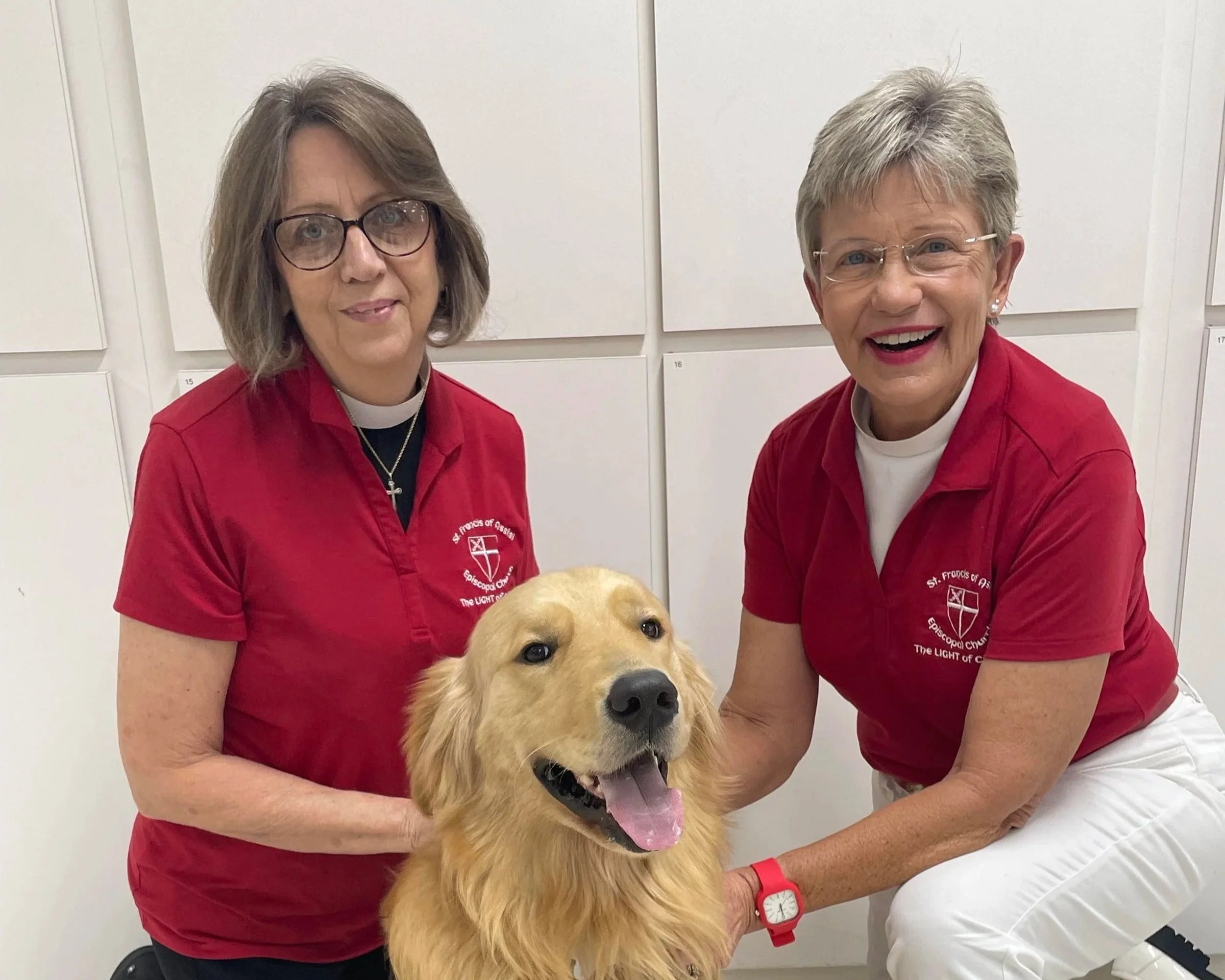 Two women wearing red shirts, one with glasses and the other with short gray hair, smiling and posing with a golden retriever dog in a setting with white lockers in the background.